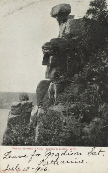 Black and white photographic postcard of the rock formation called the Great Stone Face in Devil's Lake State Park. A man is standing to the right of the "face." Trees and shrubs are growing on the rocks. The lake, bluffs and sky are in the background. The text below reads: "Great Stone Face, Devil's Lake, Wis."