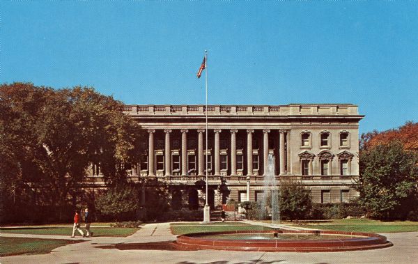 Color postcard of an exterior view of the Wisconsin State Historical Society Building, now the headquarters of the Wisconsin Historical Society. In the foreground pedestrians are walking near the fountain in the middle of the Library Mall.