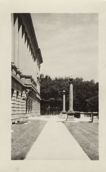Photographic postcard of view down the front sidewalk of the main entrance of the State Historical Library, now the headquarters building of the Wisconsin Historical Society. Trees are in the background.