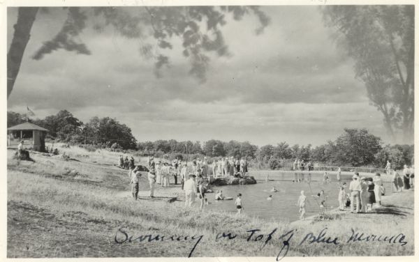 Photographic postcard view of the swimming pool on top of the mounds in Blue Mounds State Park. There are people swimming as well as standing around the sides of the pool watching. The clouds and transparent trees were added to the scene, see the Public Notes. The sender wrote on the bottom of the postcard in ink, "Swimming on Top of Blue Mounds."