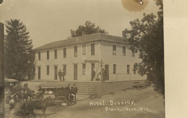 Photographic postcard of the Hotel Donnelly. Men are standing and visiting on the porch. A sign on the corner reads: "Hotel & Livery." Horse-drawn vehicles and passengers are waiting in the street at the foot of the steps. Trees are on the left and right. Below is the text: "Hotel Donnelly, Bloomington, Wis."