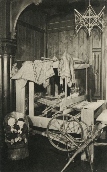Black and white postcard of an indoor scene with a loom, spinning wheel, skeiner, garments and two dolls seated in a chair at Little Norway.
