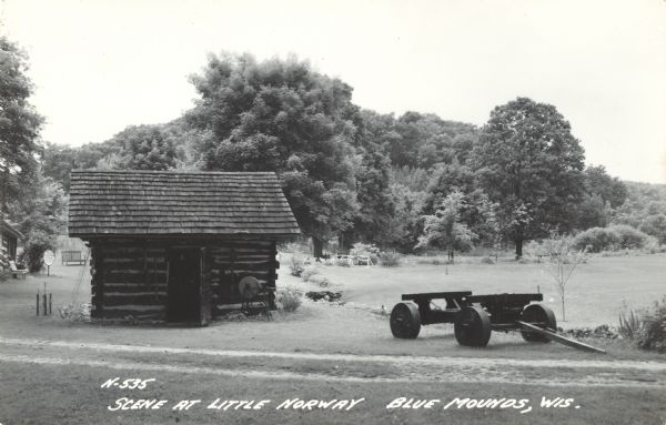 Photographic postcard of a scene at Little Norway. A log building stands to the left and the chassis of a wagon to the right. Text below reads: "Scene at Little Norway, Blue Mounds, Wis."