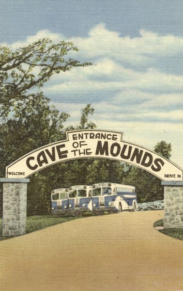 Colorized postcard of the entrance to the Cave of the Mounds. A curved sign sits on two square stone pillars. The sign reads "Welcome, Cave of the Mounds, Drive In." Three blue and white tour buses are visible through the arch as well as cars. Text on the back reads: "Parking area-for one thousand cars, Cave of the Mounds, Blue Mounds, Wisconsin. 25 miles west of Madison, Wis. on Highway 18-151."