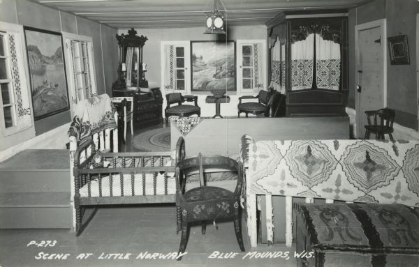 Photographic postcard of the main living area of a Norwegian dwelling at Little Norway showing a cradle, chairs, trunks, dresser and a built-in bed with curtains. Large paintings decorate the walls and a lantern hangs from the ceiling. Text below reads: "Scene at Little Norway, Blue Mounds, Wis."