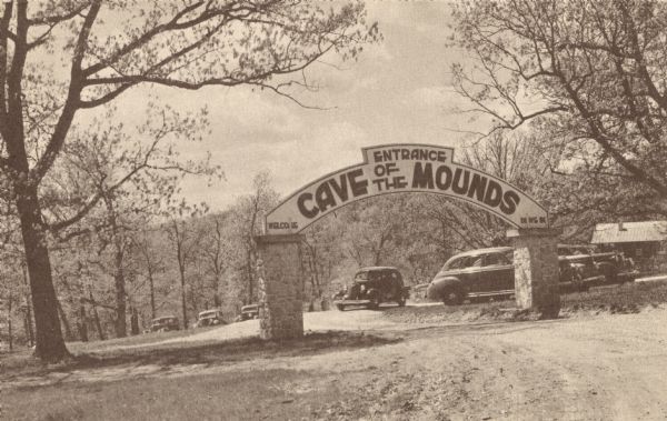 Postcard of the entrance to the Cave of the Mounds. A curved sign sits on two square stone pillars. The sign reads: "Welcome, Cave of the Mounds, Drive In." Automobiles are visible through the arch. Text on the back reads "The Oak Grove parking area at the entrance to Dave of the Mounds, Blue Mounds, Wis."