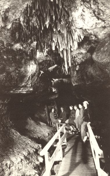Photographic postcard of Crystal Chandeliers above the Lake Bridge in the Cave of the Mounds. A group of visitors stand on a wooden walkway underneath the formations.