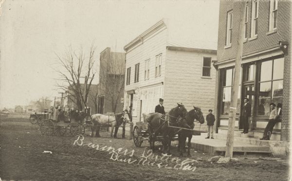 Photographic postcard of the business district in Blue River. Stores and a wooden boardwalk line the dirt street, and horse-drawn wagons wait in front of the buildings. One wagon is full of people. Several individuals are standing on the boardwalk. Handwritten text below reads: "Business District, Blue River, Wis."