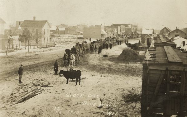 Elevated view of Stock Days. Horses and wagons loaded with livestock are lined up in a row on the snowy street. On the right is a train decorated with American flags. Railroad cars are carrying livestock. On the left is a street lined with buildings.
