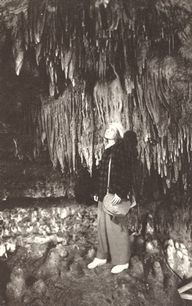 Photographic postcard of a woman viewing stalactites on the ceiling and dome shaped stalagmites on the floor of the Chapel, North Cave, in the Cave of the Mounds.