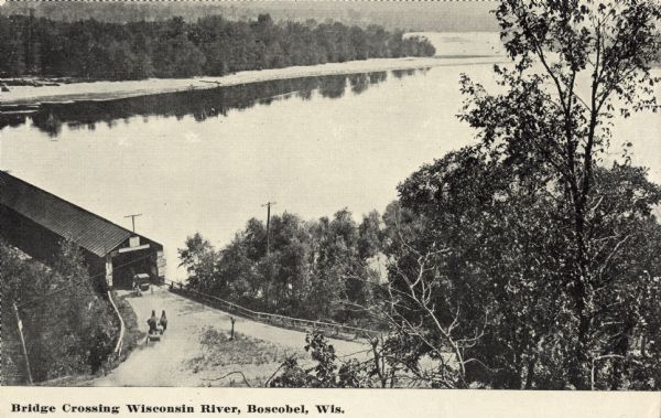 Black and white postcard of an elevated view of the Old Covered Bridge crossing the Wisconsin River. The bridge is on the left and the river is on the right. Two horse-drawn vehicles are driving onto the bridge. The text below reads: "Bridge Crossing Wisconsin River, Boscobel, Wis."