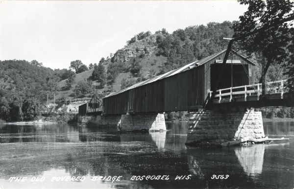 Photographic postcard of the Old Covered Bridge across the Wisconsin River at Boscobel. Bluffs and trees are in the background. Caption reads: "The Old Covered Bridge, Boscobel, Wis."