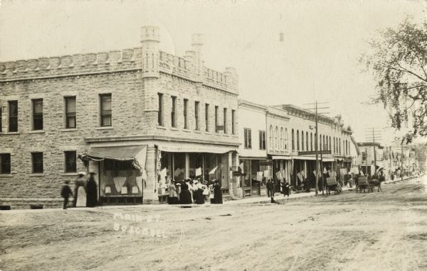 Photographic postcard of Main Street. Many people can be seen on the sidewalks and flags decorate the buildings. The street is not paved and several horse-drawn buggies are parked at the curb. Caption reads: "Main St., Boscobel, Wis."