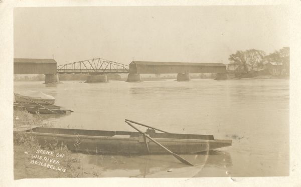View of three boats on a shoreline with the covered bridge near Boscobel spanning the Wisconsin River in the distance. The text in the lower left corner reads "Scene on Wis. river, Boscobel, Wis."