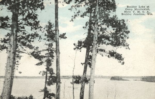 Colorized postcard of Boulder Lake. Pine trees are in the foreground and islands are below in the background. Caption reads: "Boulder Lake at Camp Manitowish, State Y.M.C.A., Boulder Junction, Wis."