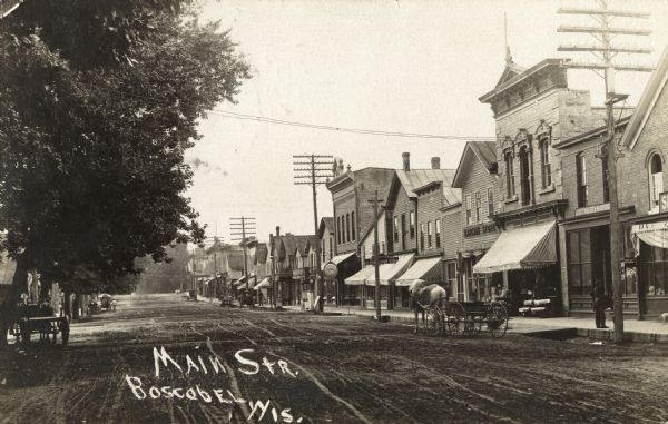 Photographic postcard of Main Street. Buildings and trees line the street. Horse-drawn vehicles and pedestrians are on the street and the sidewalks. Text below reads: "Main Str., Boscobel, Wis."