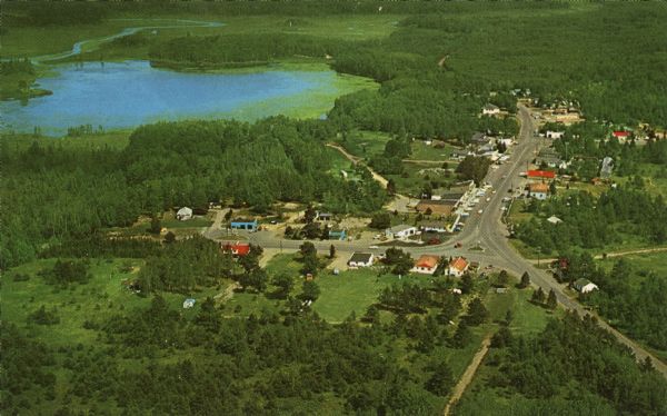 Color postcard of an aerial view of town. There is a lake in the background in the upper left.