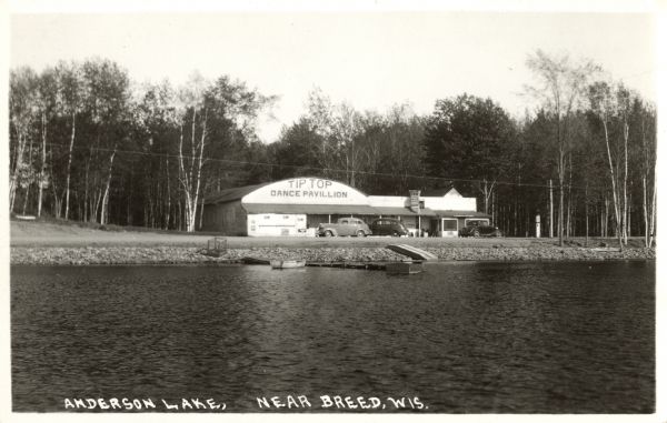 Photographic postcard of the Tip Top Dance Pavillion on Anderson Lake. The view from the lake shows several automobiles parked outside the pavillion. The shore is rocky, and there is a pier with one boat tied up. Trees are in the background. Text below reads: "Anderson Lake, Near Breed, Wis."