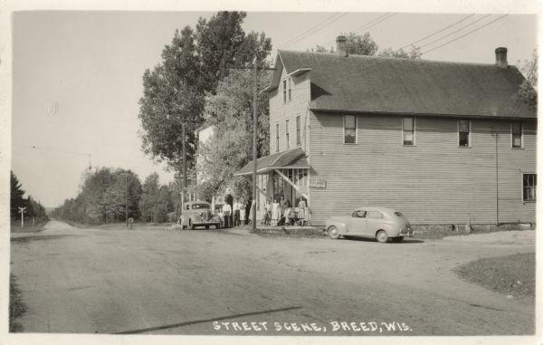Photographic postcard down road toward a building on the right. Signs indicate that the Post Office and Fire Marshall can be found within. Two automobiles are parked outside. A group of people and a dog are on the porch. Another group is posed around the Mobil gas pump. Caption reads: "Street Scene, Breed, Wis."