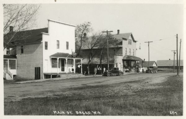 Photographic postcard view toward buildings along the left on Main Street. A tavern is next to a general store with a gas pump on the corner. Two automobiles are parked outside. One man sits on the porch and another is by his car. A group of people stand between the buildings. Caption reads: "Main Street, Breed, Wis."