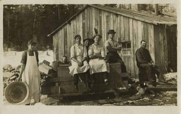 Photographic postcard of a group of workers, all men, and one boy, posing outside of a wooden building. Three of the workers wear white aprons. Four of the men sit on a sled, one man sits on a barrel, and another man is standing holding a washtub. There are trees and snow on the ground.