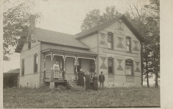 Photographic postcard of a group of adults posing in front of a brick house. The house has a patterned design on the exterior. Below the peak of the roof, the date 1902, created with bricks, can be seen. Trees surround the house.