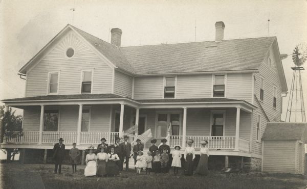 Photographic postcard of group of people posing in front of a two-story wooden home with a large porch on which American Flags are displayed. A windmill stands behind the house on the right. A small toy car is on the lawn in front of the porch.