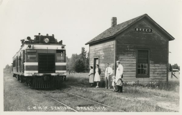 Photographic postcard of a train stopped at the Chicago & North West railroad station. A small group of people stand in the grass waiting beside the tracks. Caption reads: "C. & N.W. Station, Breed, Wis."