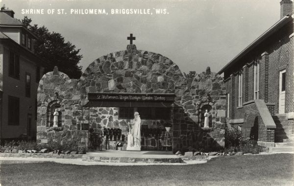 Photographic postcard of the Shrine of Saint Philomena. It is a large stone structure with a marquee over the statue in the center that reads: "St. Philomena, Virgin Martyr, Wonder Worker." In two niches on each side smaller statues are displayed. A church building is on the right and a house is on the left. Caption reads: "Shrine of St. Philomena, Briggsville, Wis."