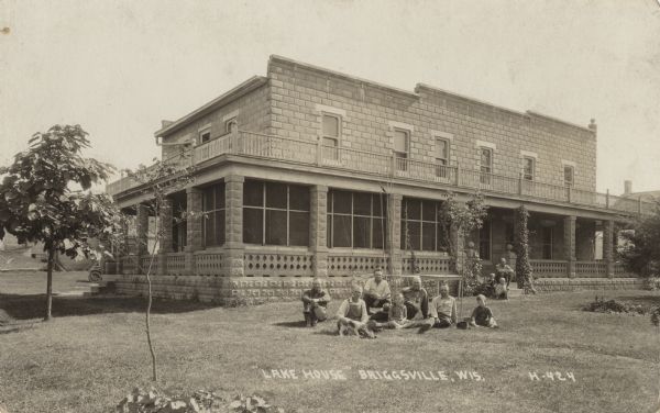 Photographic postcard view of a group of men, children and a dog posing in front of Lake House. Another man with two children are sitting on the porch steps. The house is built of stone with an extensive wrap-around porch. Above the porch is a balcony. Caption reads: "'Lake House' Briggsville, Wis."