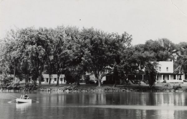 Photographic postcard view across water towards the Pheasant Inn on Lake Mason. Buildings are along the shoreline behind large trees. Two people are in a rowboat in the foreground. Text on the back reads: "'Pheasant Inn' Lake Mason, Briggsville, Wis. Modern Heated Motel - Fine Food - Bar, Excellent Fishing and Hunting."