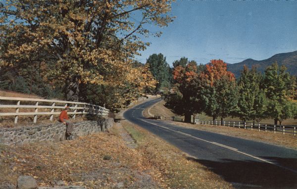 Color postcard of a man sitting on a stone wall along a road while enjoying a country scene. The road curves through the hills and the trees are starting to show autumn color. White fences line both sides.