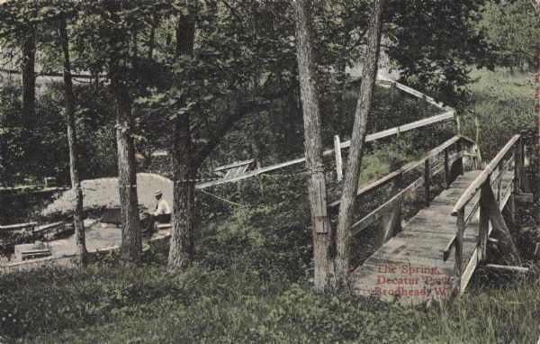 Colorized postcard view of The Spring in Decatur Park. A man is sitting near the spring. There is a bridge on the right, and many trees. Text in red in the lower right corner reads: "The Spring, Decatur Park, Brodhead, Wis."