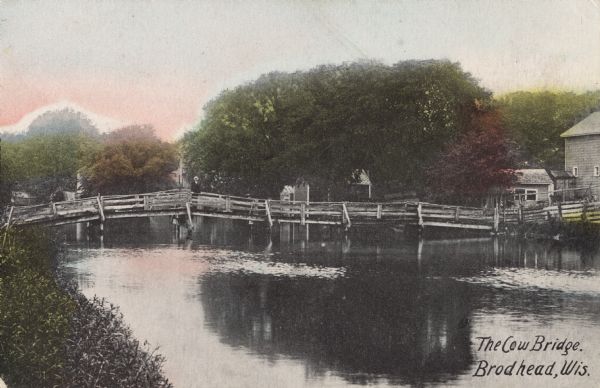 Colorized postcard view of the Cow Bridge. A farm is on the shoreline on the right. A man is standing on the bridge. Text in the lower right corner reads: "The Cow Bridge, Brodhead, Wis."