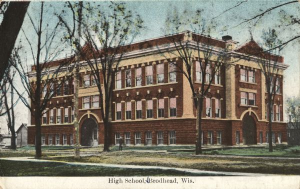 Colorized postcard of the Brodhead High School. A student is standing on the lawn in front, with many trees surrounding the building. Caption reads: "High School, Brodhead, Wis."