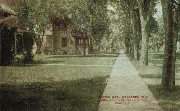 Colorized postcard view of a neighborhood on Clinton Avenue. View down sidewalk of the tree-lined street of homes with large front lawns. Caption reads: "Clinton Ave., Brodhead, Wis."