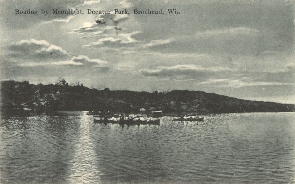 Colorized postcard view across water towards people in boats on the lake in Decatur Park. It is evening and the moon is shining on the lake. Caption reads: "Boating by Moonlight, Decatur Park, Brodhead, Wis."