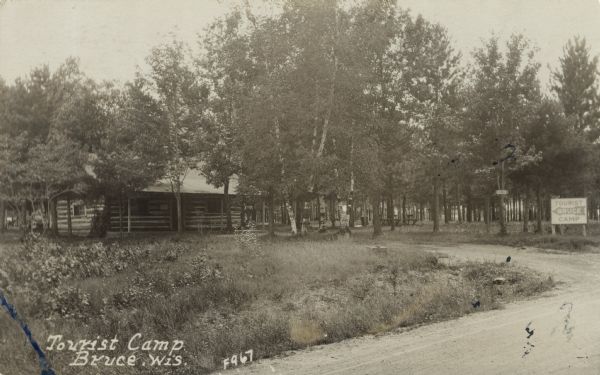 Photographic postcard view of the Bruce Tourist Camp. A dirt road curves into the camp, with a large sign at the entrance, and cabins, picnic tables and many trees. Caption reads: "Tourist Camp, Bruce, Wis."