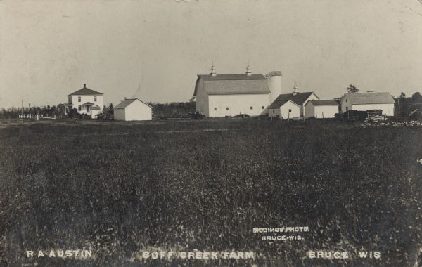Photographic postcard view of Buff Creek Farm. The farmhouse is on the left and the barn and outbuildings are on the right. A man and an automobile are near the barn. Caption reads: "R.A. Austin, Buff Creek Farm, Bruce, Wis."