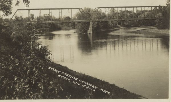 Photographic postcard view from shoreline of a bridge over the Chippewa River. Caption reads: "Bridge Crossing Chippewa River, Bruce, Wis."