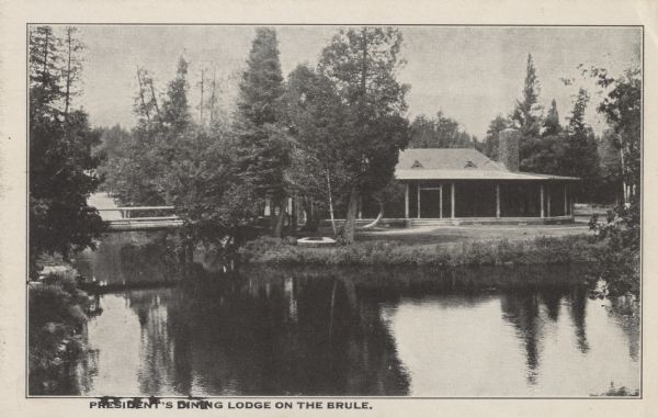 Black and white postcard view of the President's Dining Lodge on the Brule River surrounded by trees. A bridge is on the left. Caption reads: "President's Dining Lodge on the Brule."