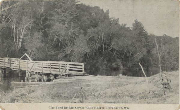 Black and white postcard view of the wooden Ford Bridge. Trees on a hill are on the far shoreline. Caption reads: "The Ford Bridge Across Willow River, Burkhardt, Wis."