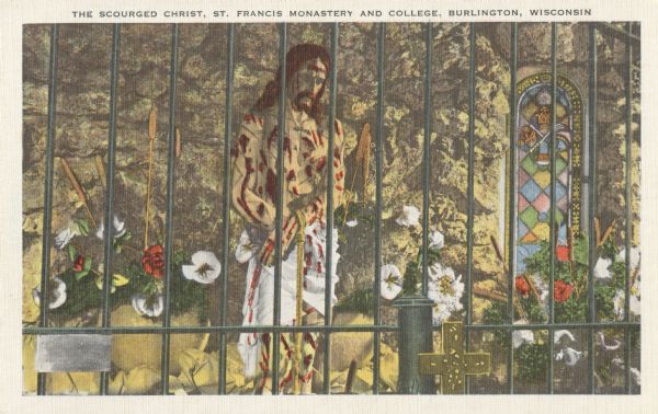 Colorized postcard of the Scourged Christ, St. Francis Monastery and College. The statue is behind bars, in front of a stone wall. On the right is a stained glass window, flowers and cattails standing in vases, and a cross in the foreground. Text at top reads: "The Scourged Christ, St. Francis Monastery and College, Burlington, Wisconsin."