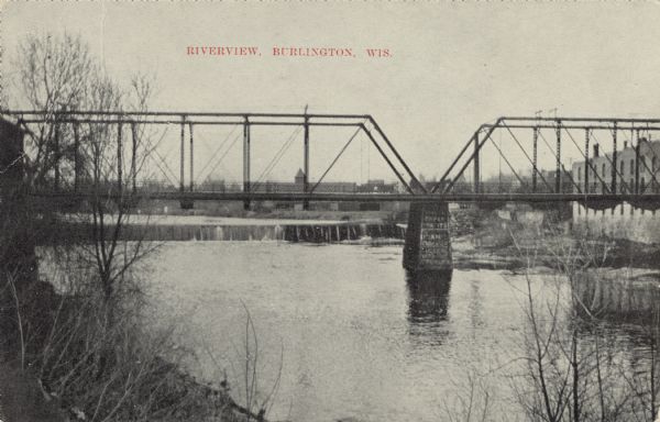 Black and white postcard of a river view of the Fox River as it runs through the city of Burlington. In the foreground is a railroad bridge. Just behind the bridge is a spillway over a dam, and the city in the background. Caption reads: "Riverview, Burlington, Wis."