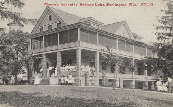 Black and white postcard of Moore's Lakeside Resort on Brown's Lake. Staff and/or visitors are posing on the front porch and on the lawn. The wrap-around balcony above the porch is screened. In the background on the left there appears to be a water tower. Text above reads: "Moore's Lakeside, Brown's Lake, Burlington, Wis."