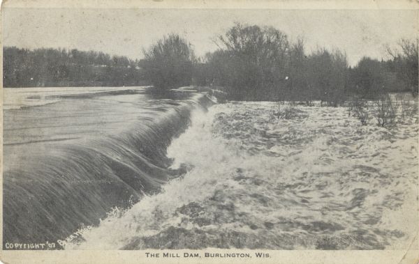 Black and white postcard of the Mill Dam. The water is running over the dam from the left and becoming rapids on the right. There is a wooded shoreline on the opposite side. Text at bottom reads: "Copyright '07 by H.A. Wood" and "The Mill Dam, Burlington, Wis."