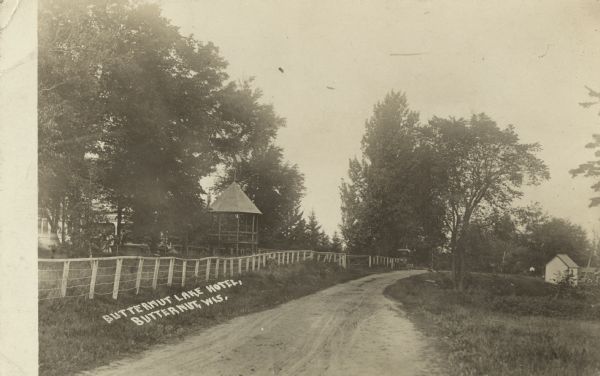 Photographic postcard view of the Butternut Lake Hotel. The road, fence, trees and screened gazebo are in the center. A man is standing among the trees near the gazebo. The Hotel is partially hidden by trees on the left. Handwritten on the negative is the text: "Butternut Lake Hotel, Butternut, Wis."
