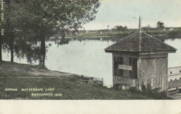 Colorized postcard of Butternut Lake in the spring, with a pier and latticed boathouse. The opposite shore has trees and foliage. Text below reads: "Spring Butternut Lake, Butternut, Wis."