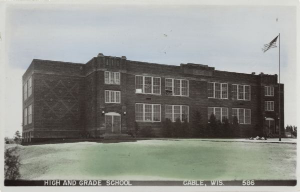 Tinted photographic postcard of Cable's High and Grade School building. A flag flies on the right, and a group of children are looking out of a second story window. Caption reads: "High and Grade School, Cable, Wis."