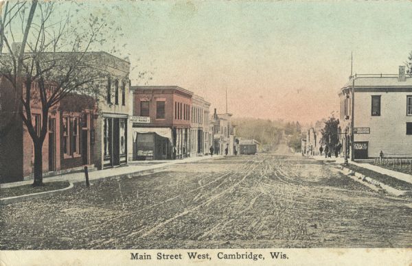 Colorized postcard of Main Street West. The street is unpaved and covered with ruts. Sidewalks line both sides, and pedestrians are walking between the storefronts. Text at foot reads: "Main Street West, Cambridge, Wis."